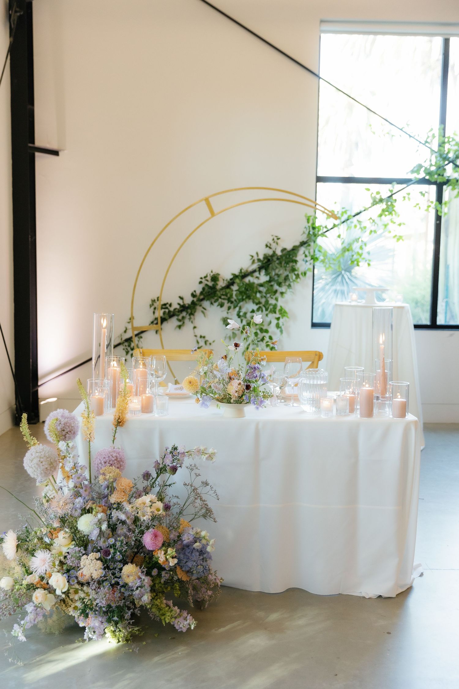 Elegant wedding table setup with gold arch, white tablecloth, candles and fresh floral arrangements in natural light.