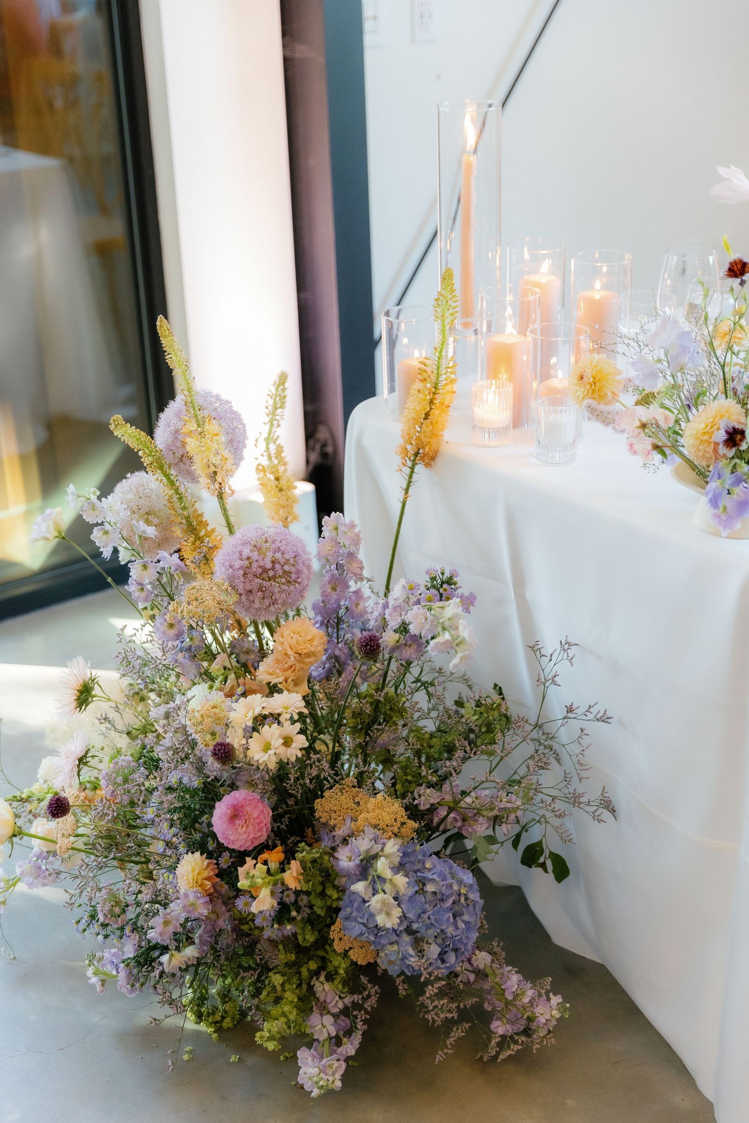 Delicate pastel floral arrangement with wildflowers and candles on a white tablecloth.