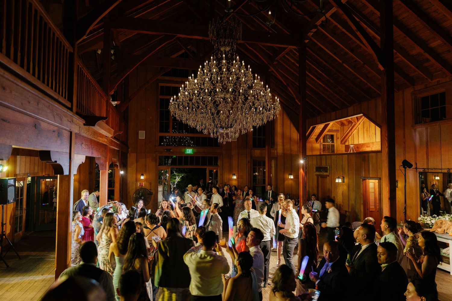 Elegant wedding reception in a rustic wooden barn venue with sparkling chandeliers and dancing guests on the dance floor.