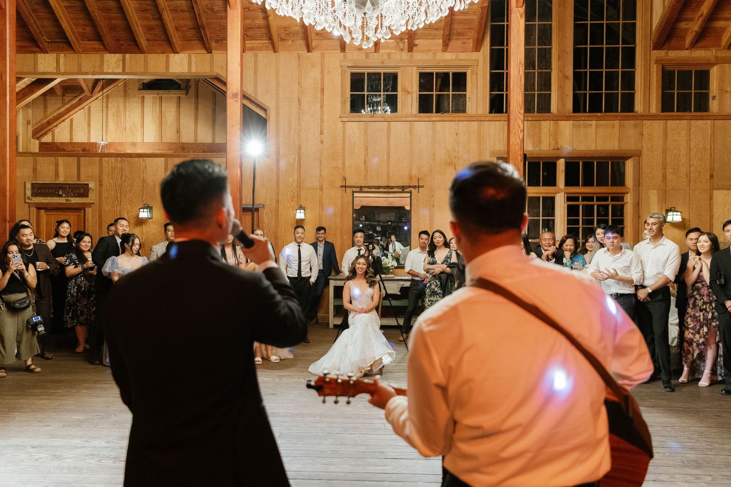 A wedding reception dance scene inside a rustic wooden barn venue with chandelier lighting.