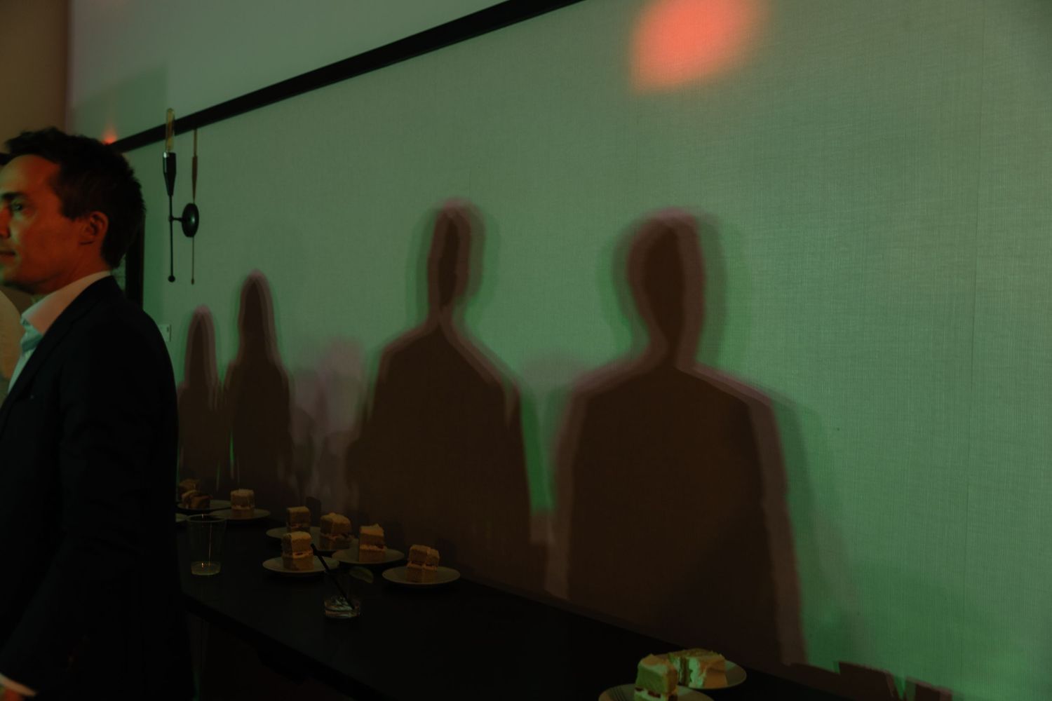Silhouettes of people cast dramatic shadows on a green-tinted wall in a dimly lit indoor setting.