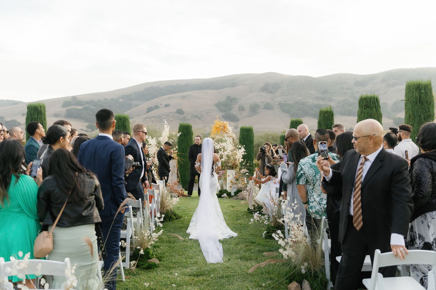 Wedding guests gather outdoors on a grassy hillside overlooking scenic mountain views during an elegant celebration.
