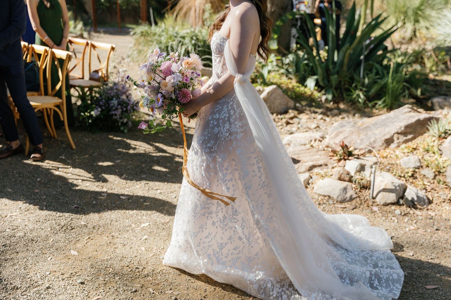 Elegant bride in a flowing white lace gown poses gracefully in a sun-dappled garden setting.