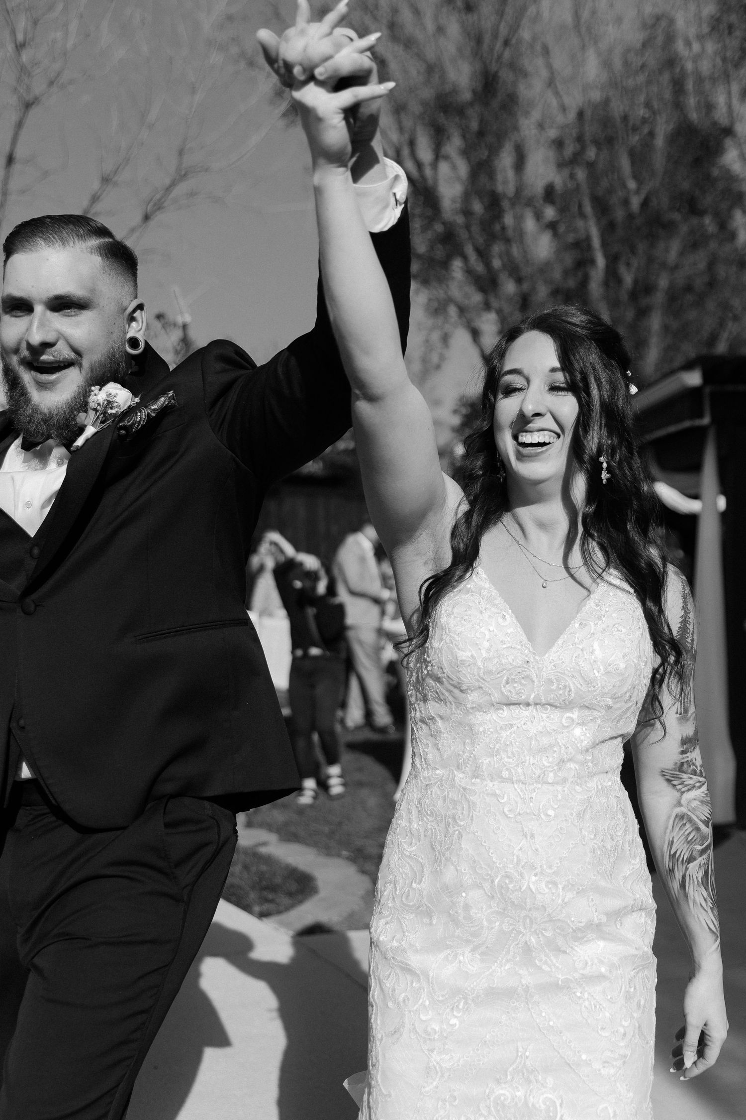 A joyful wedding couple celebrates outdoors, with the bride in white and groom in black suit raising their arms in celebration.