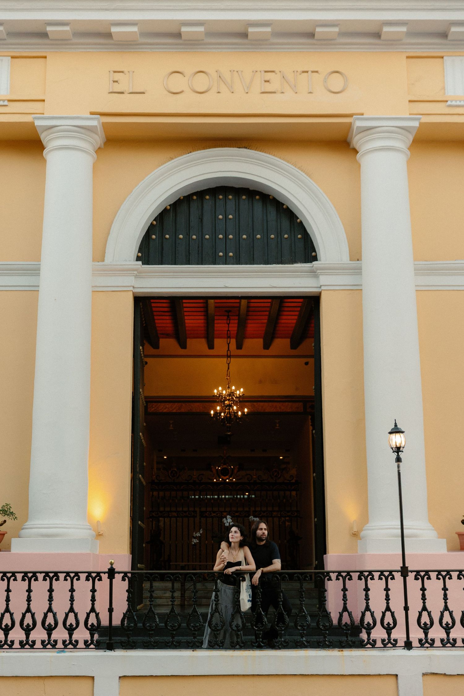 Historic yellow building entrance 'El Convento' with white columns and ornate wrought iron railing illuminated at dusk.