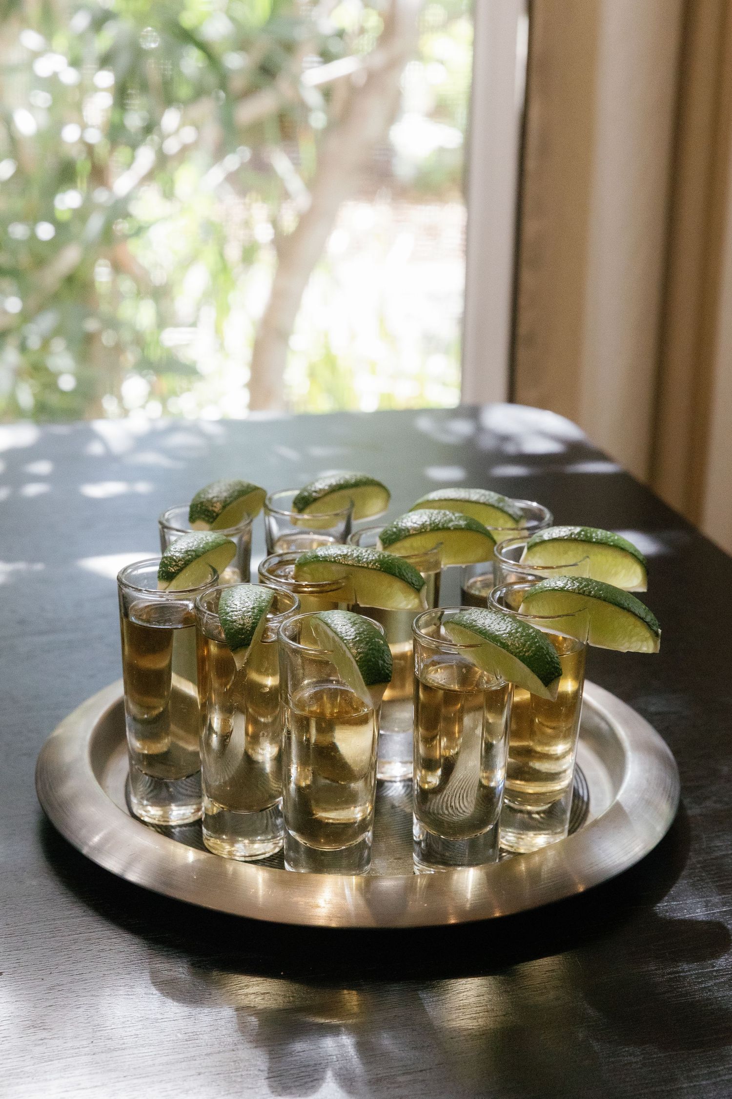 A sequence of four shots showing tequila shots with lime wedges arranged on a silver serving tray near a window.