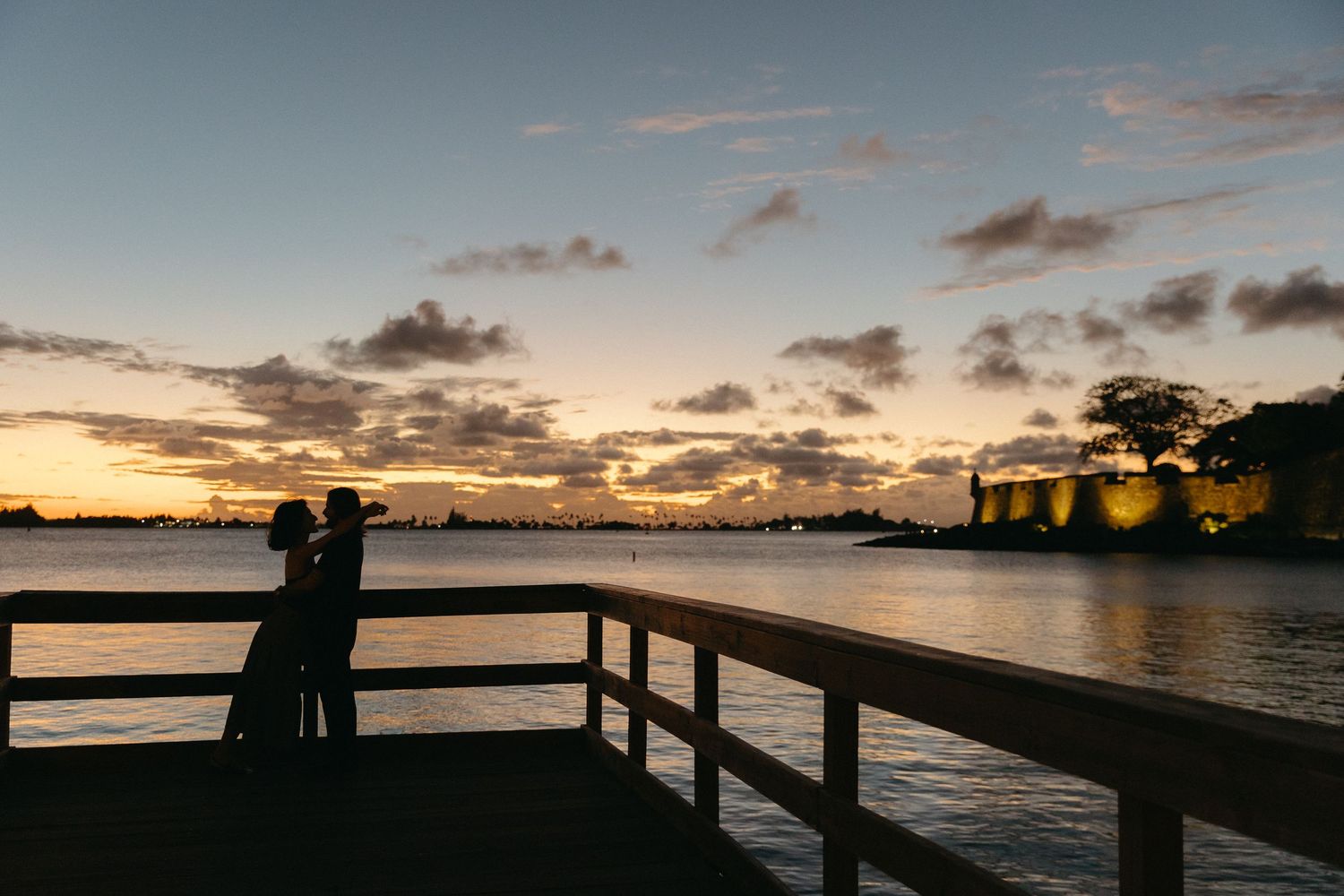A series of silhouetted figures stand on a pier watching a dramatic sunset over a calm ocean.