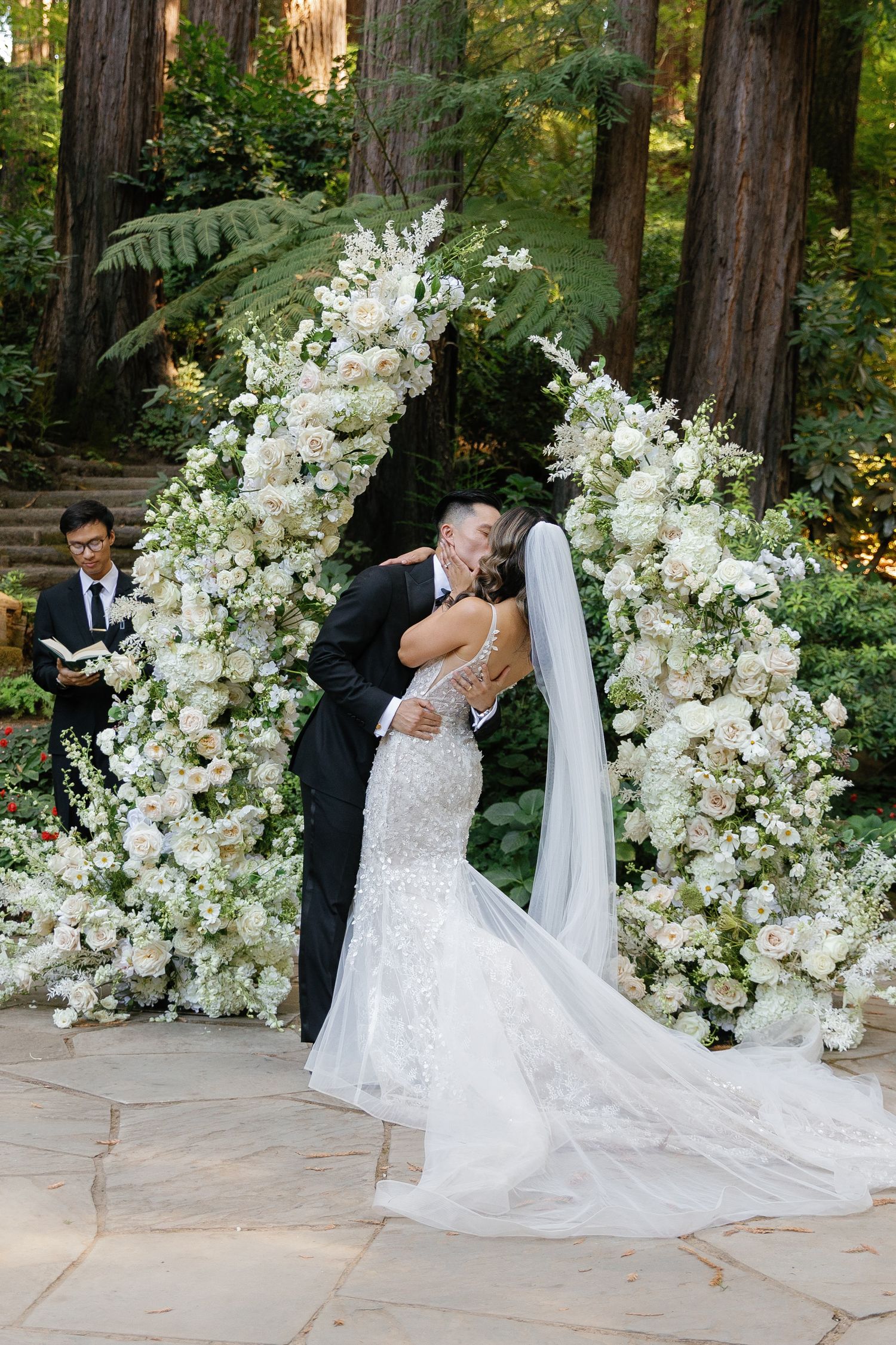 A romantic wedding ceremony takes place under a stunning white floral arch in a serene forest setting.