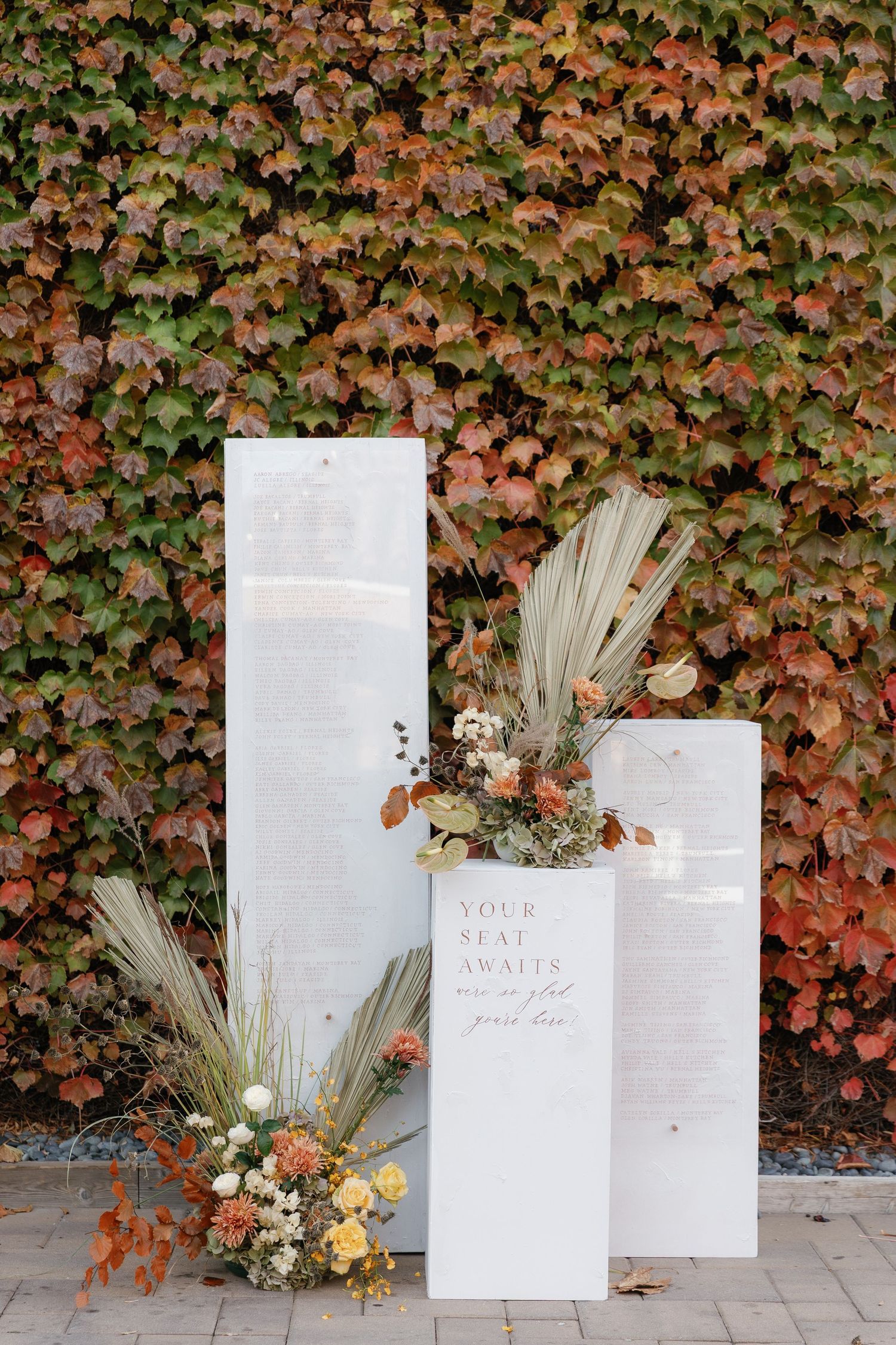 White paper luminaries with dried flowers and grasses arranged against an ivy-covered wall create an elegant autumn display.