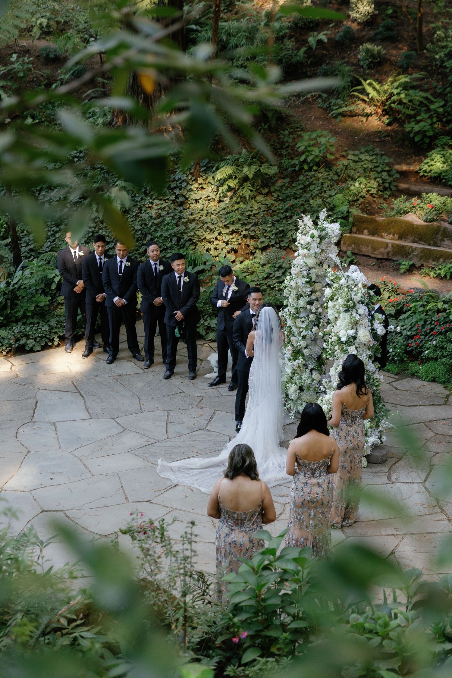 Wedding ceremony in a lush garden setting with stone pathways and white floral arrangements.