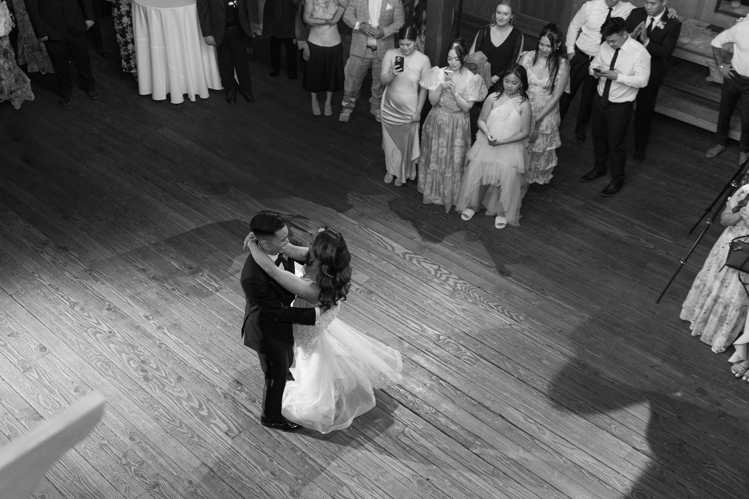 A couple shares their first dance as newlyweds on a wooden dance floor while wedding guests watch from the sidelines.