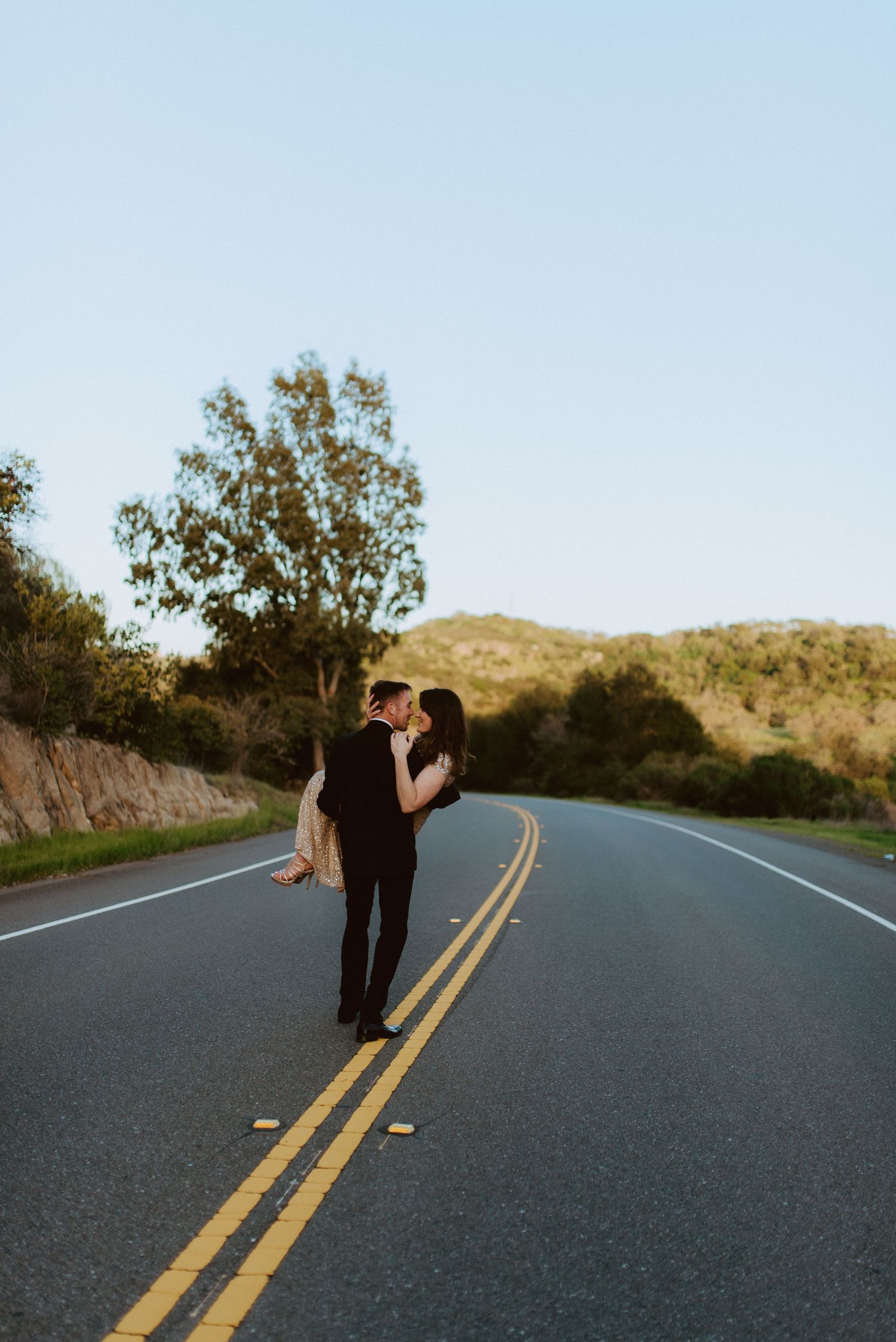 A couple embraces while standing in the middle of an empty mountain road during golden hour.