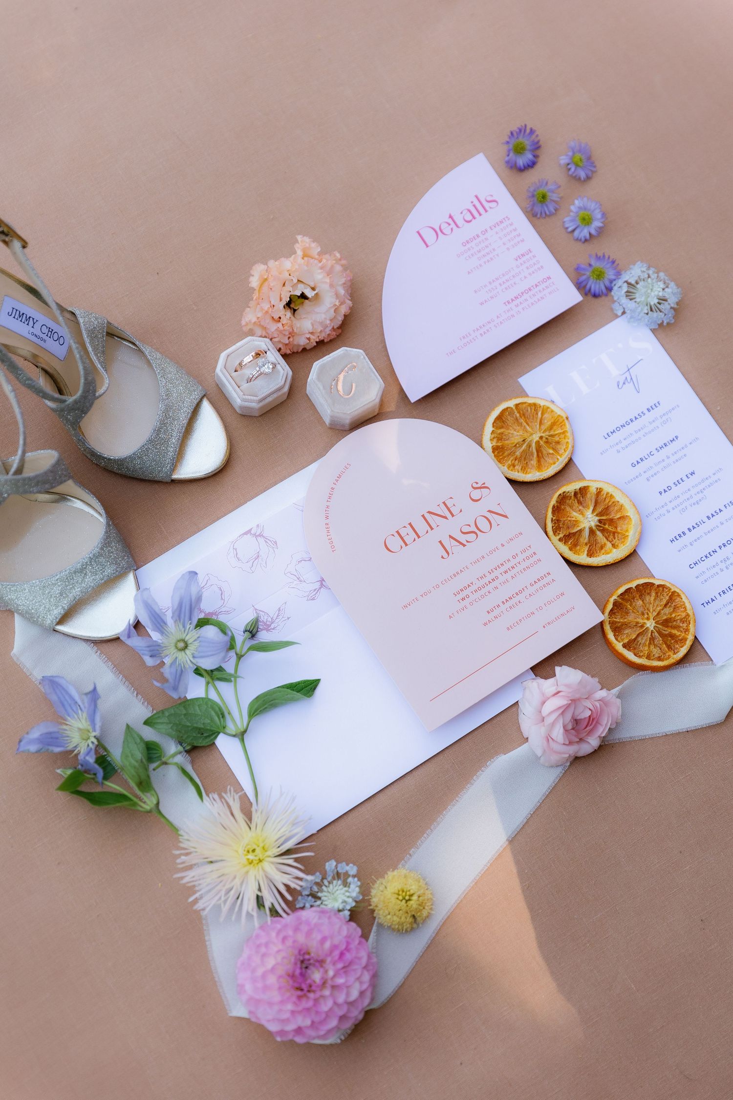 Wedding stationery flat lay with delicate pink flowers, dried orange slices, and elegant invitation cards on beige background.