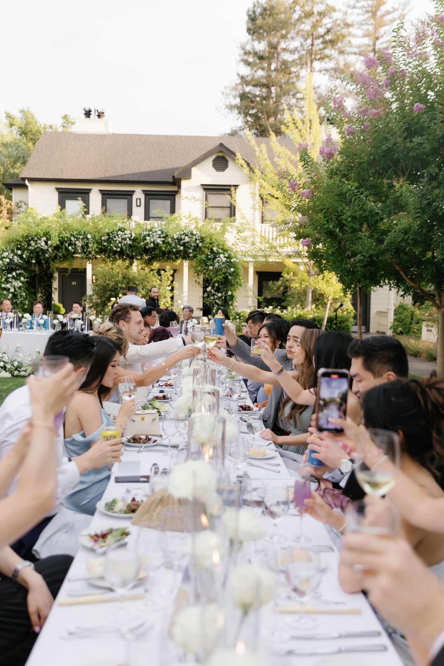 Long outdoor dinner table filled with guests raising glasses in a toast during golden hour at a garden party.