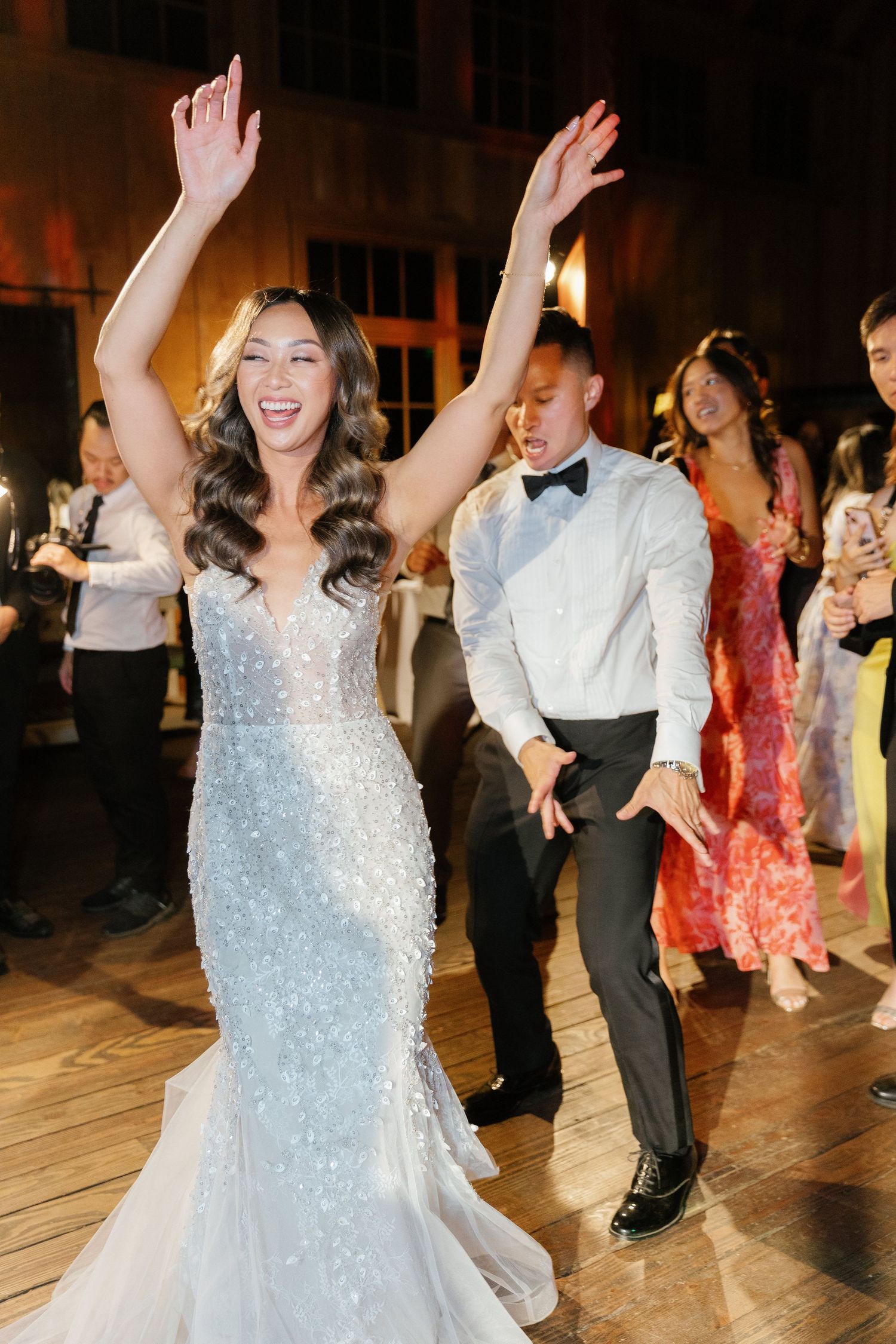 Bride in sparkly white dress dances joyfully on wooden dance floor during wedding reception celebration.