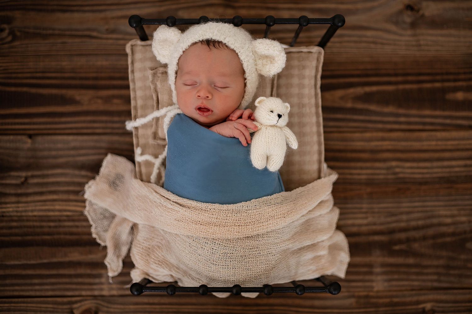 Sleeping newborn wrapped in blue swaddle rests in wooden drawer with small teddy bear and white accessories.