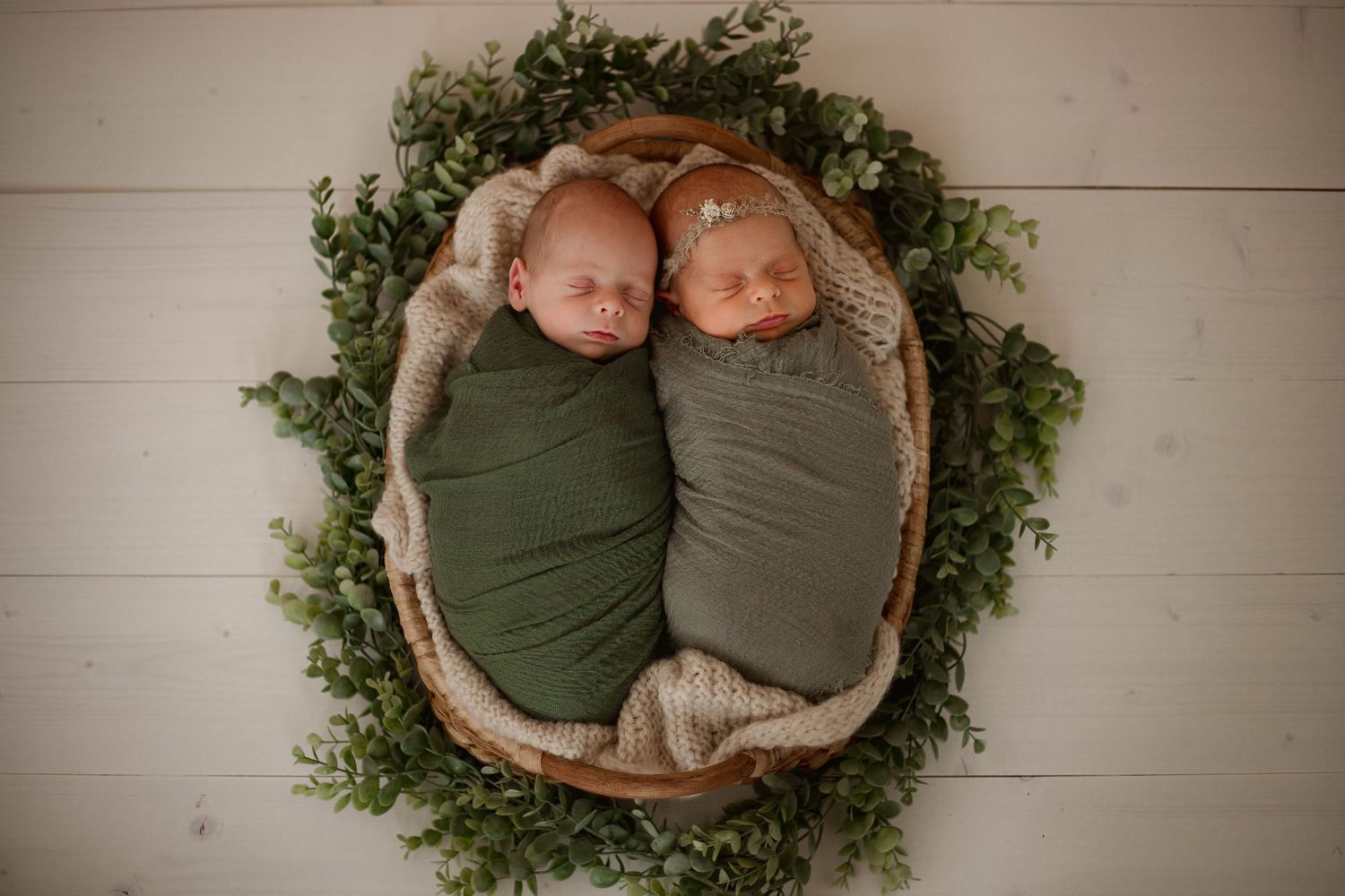 Two newborn twins swaddled in green wraps sleeping in a wooden bowl with greenery.