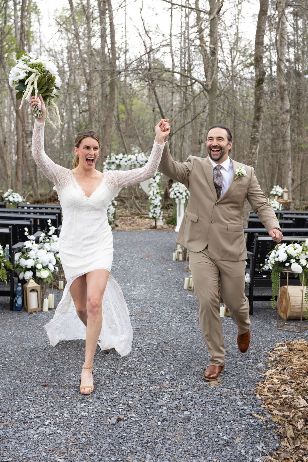 Joyful newlyweds celebrate walking down the aisle, bride in white dress holding bouquet, groom in tan suit, outdoor wedding.