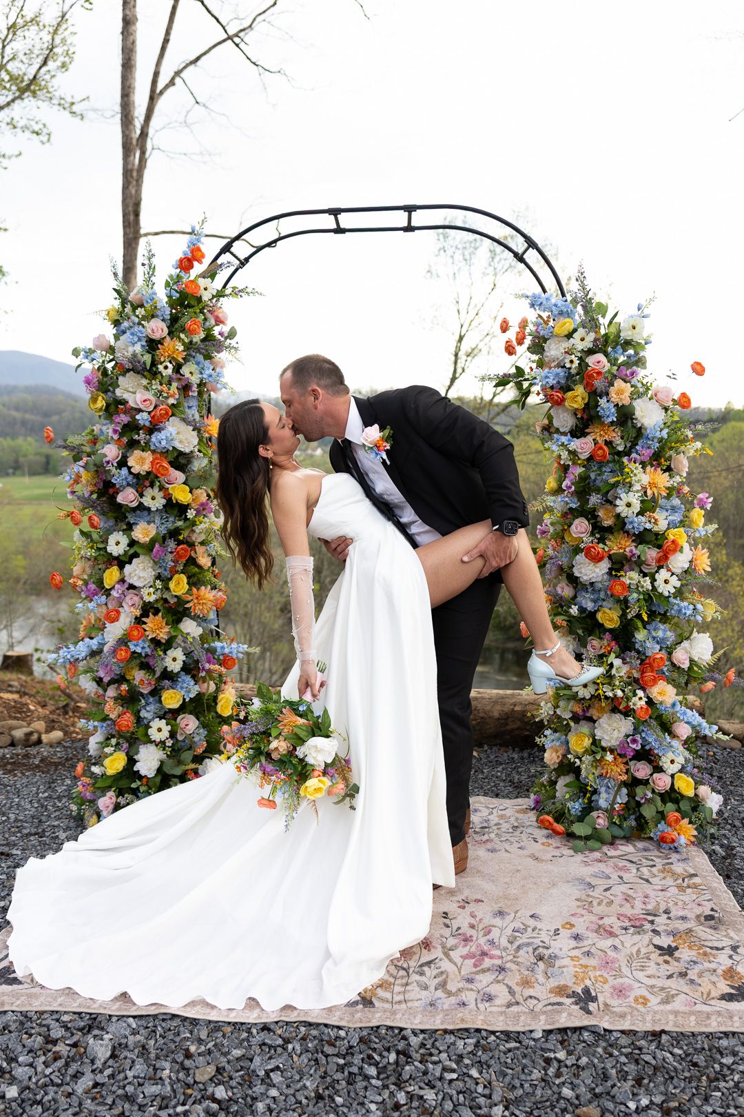 Bride and groom share a romantic dip kiss under a colorful floral arch with wildflowers at an outdoor wedding ceremony.
