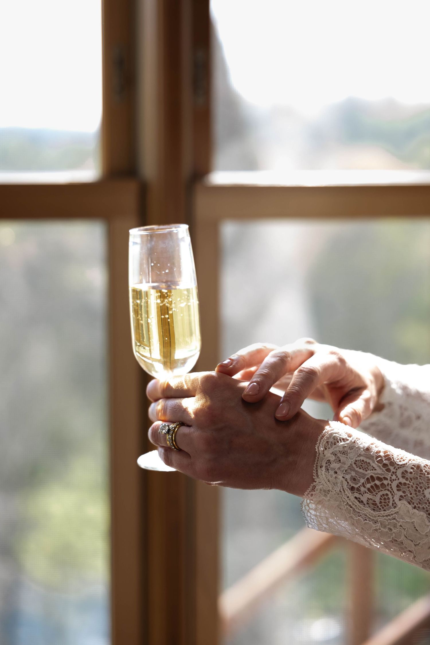 Two hands holding a champagne flute near a sunlit window, with a wedding ring visible on one finger.