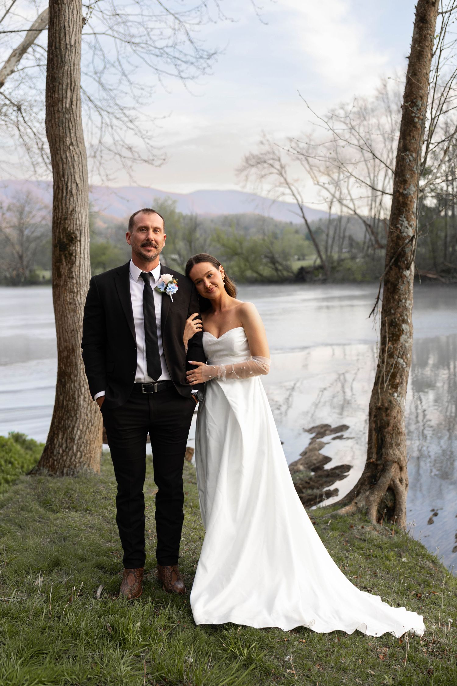 Bride and groom pose by a misty river with mountain views, she in a white gown, he in a black suit.