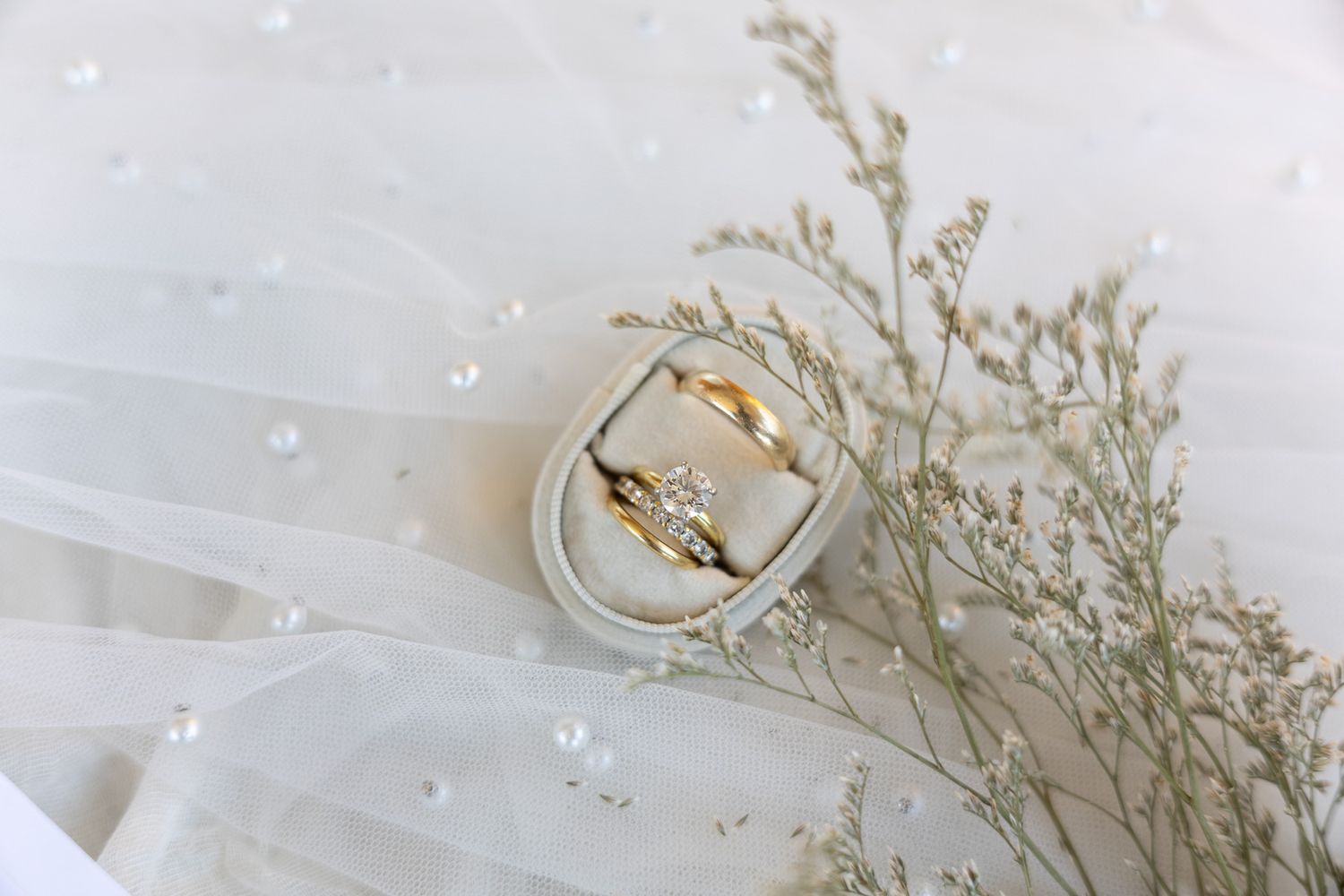 Gold wedding rings in a white velvet box on a bridal veil with pearls and dried flowers.