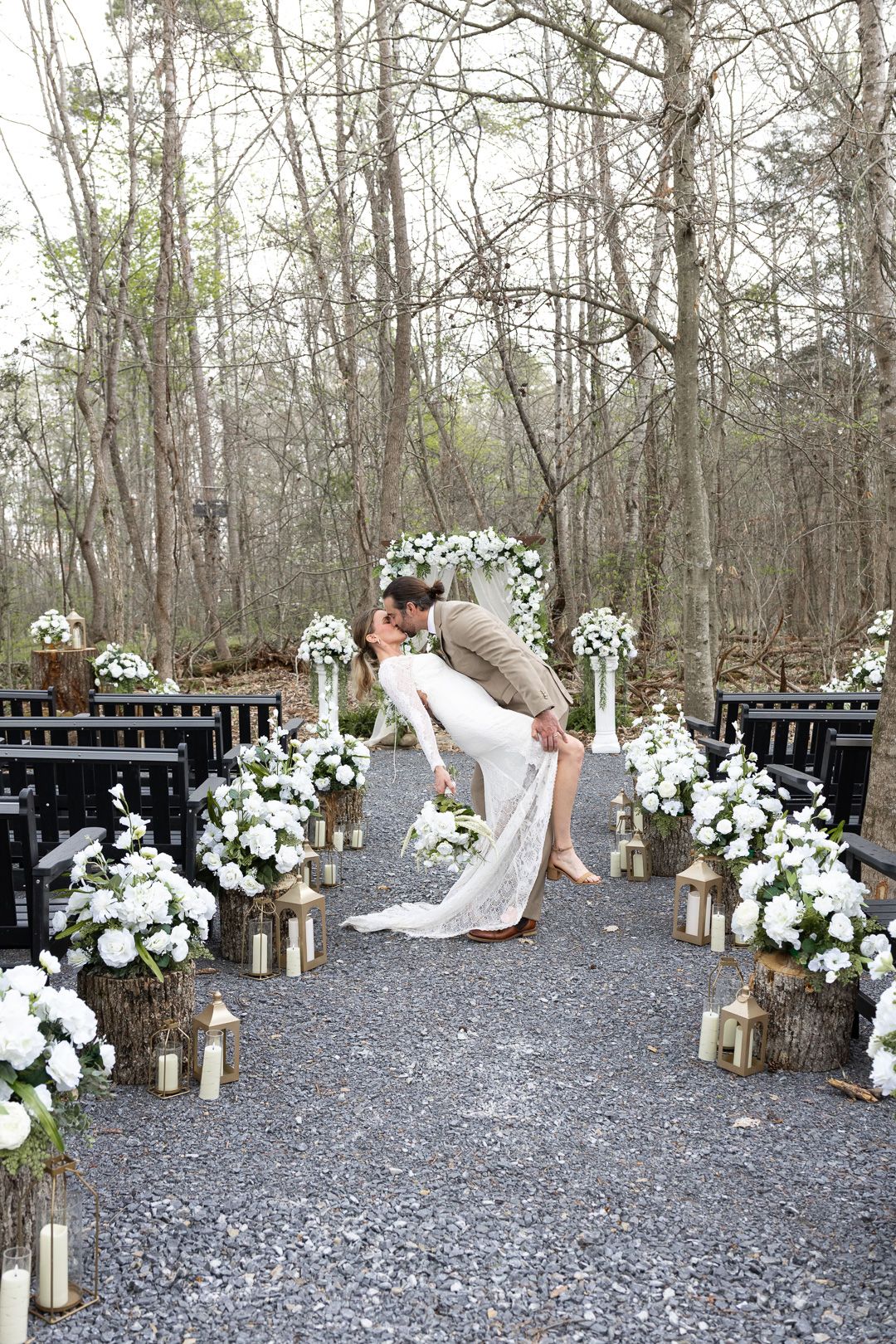 Bride and groom share a romantic dip kiss at an outdoor woodland wedding ceremony with white floral arrangements.