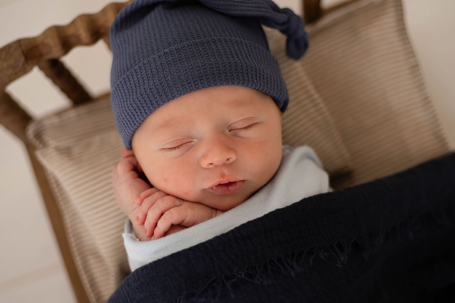 Peaceful newborn baby wearing a navy blue knit hat sleeps soundly against a dark blanket.