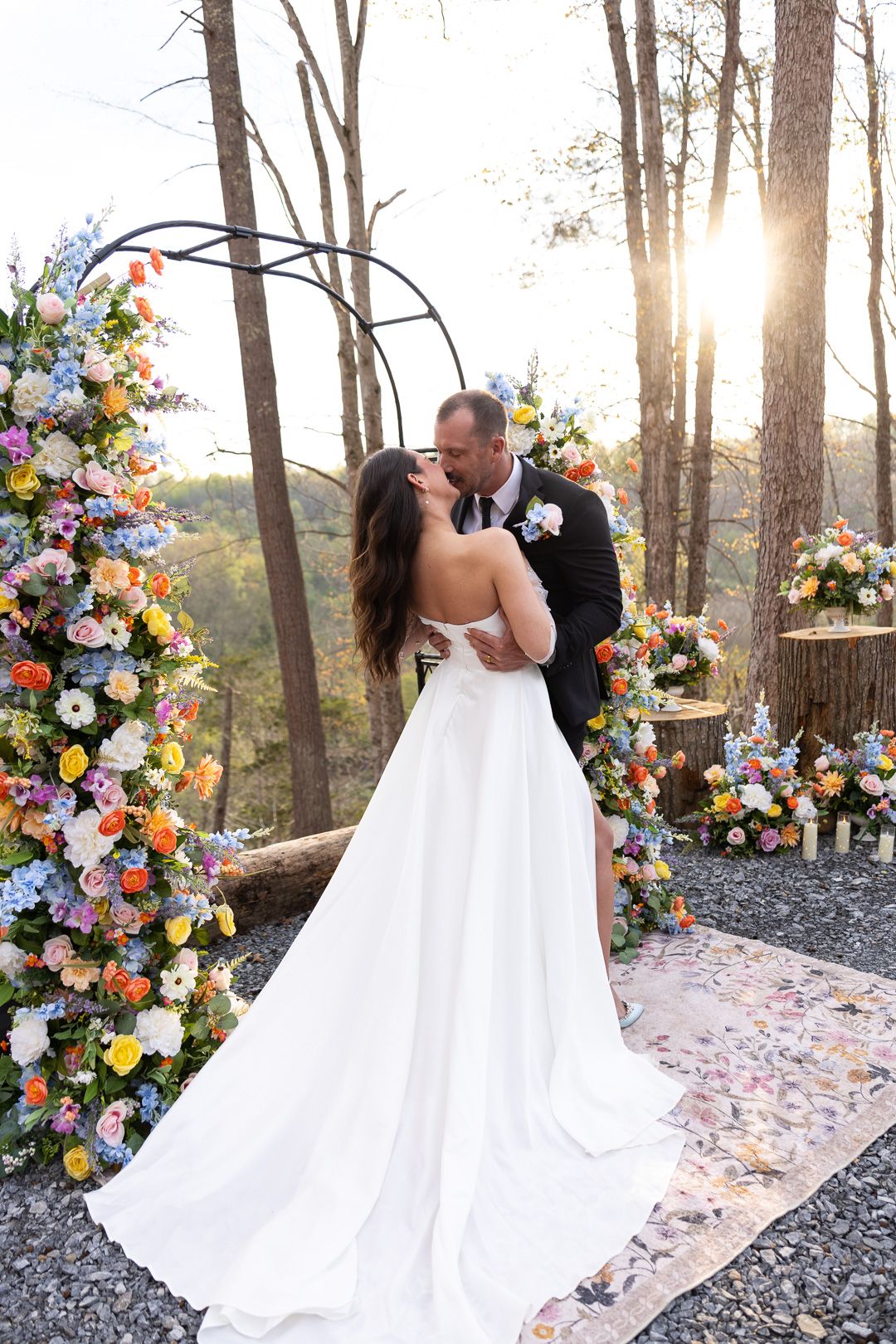 Bride and groom share a kiss under a circular floral arch at golden hour in a forest setting.