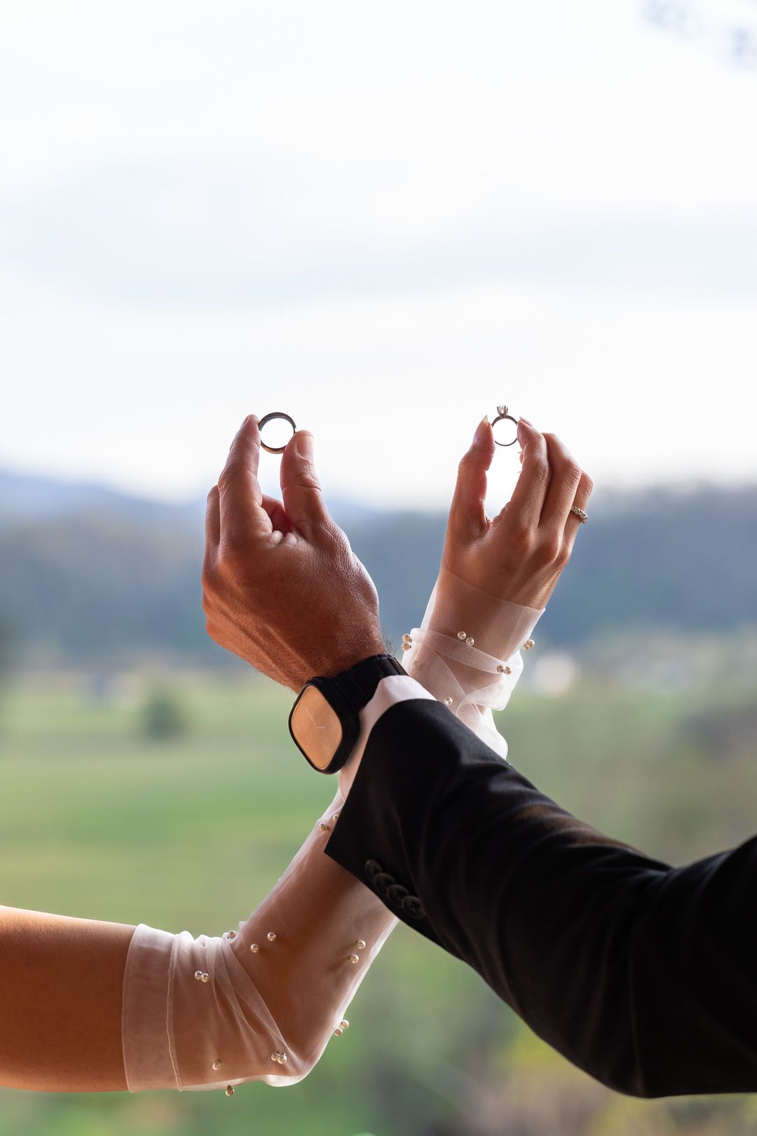 Bride and groom hold up their wedding rings together against a scenic green countryside backdrop on their wedding day.