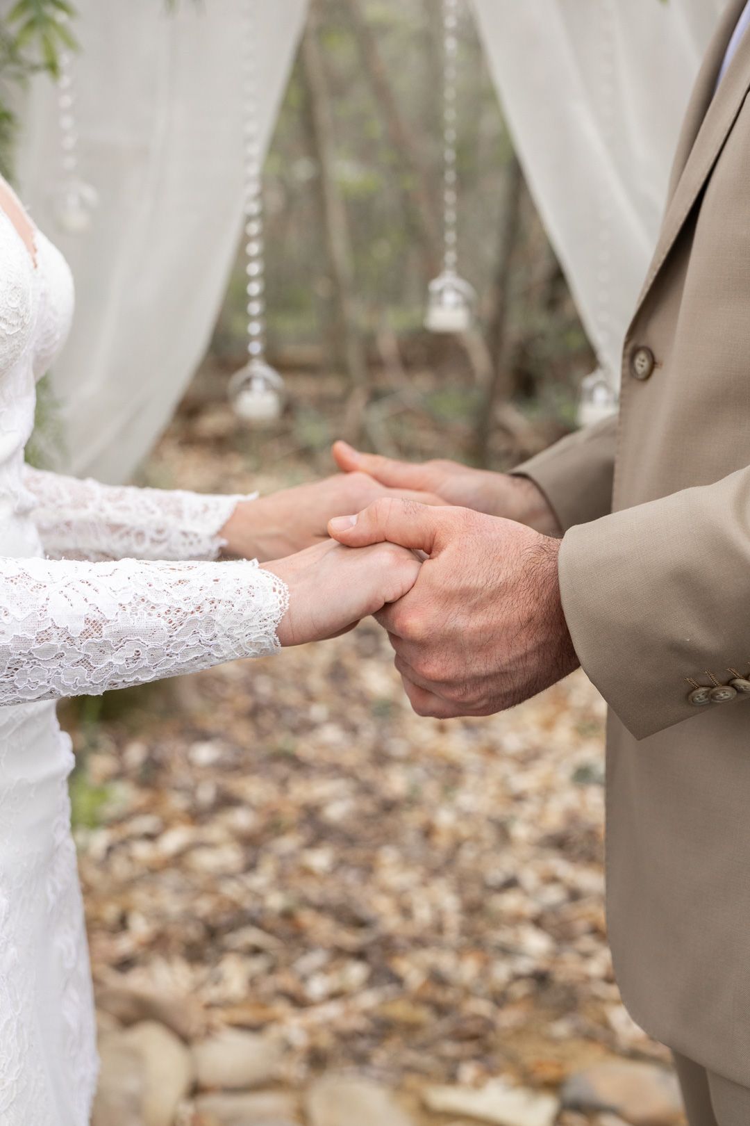Bride and groom exchange rings at an outdoor wedding ceremony, holding hands beneath a draped floral arch.