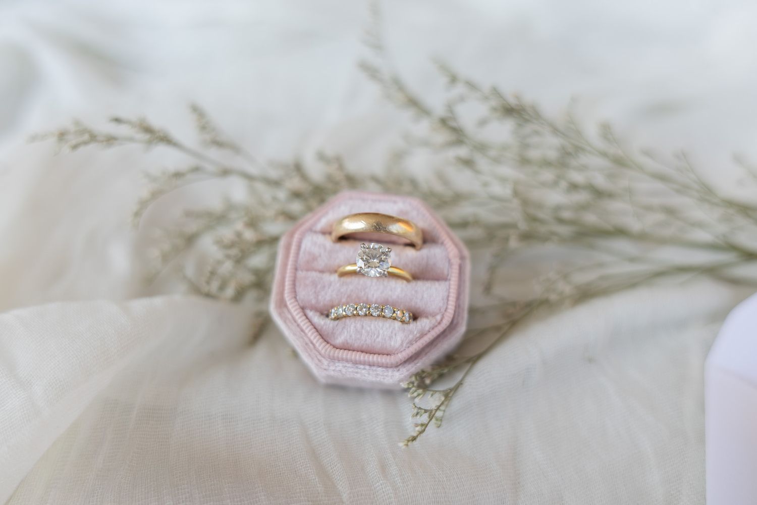 Gold wedding rings with diamond accents displayed in a pink velvet ring box surrounded by dried flowers.
