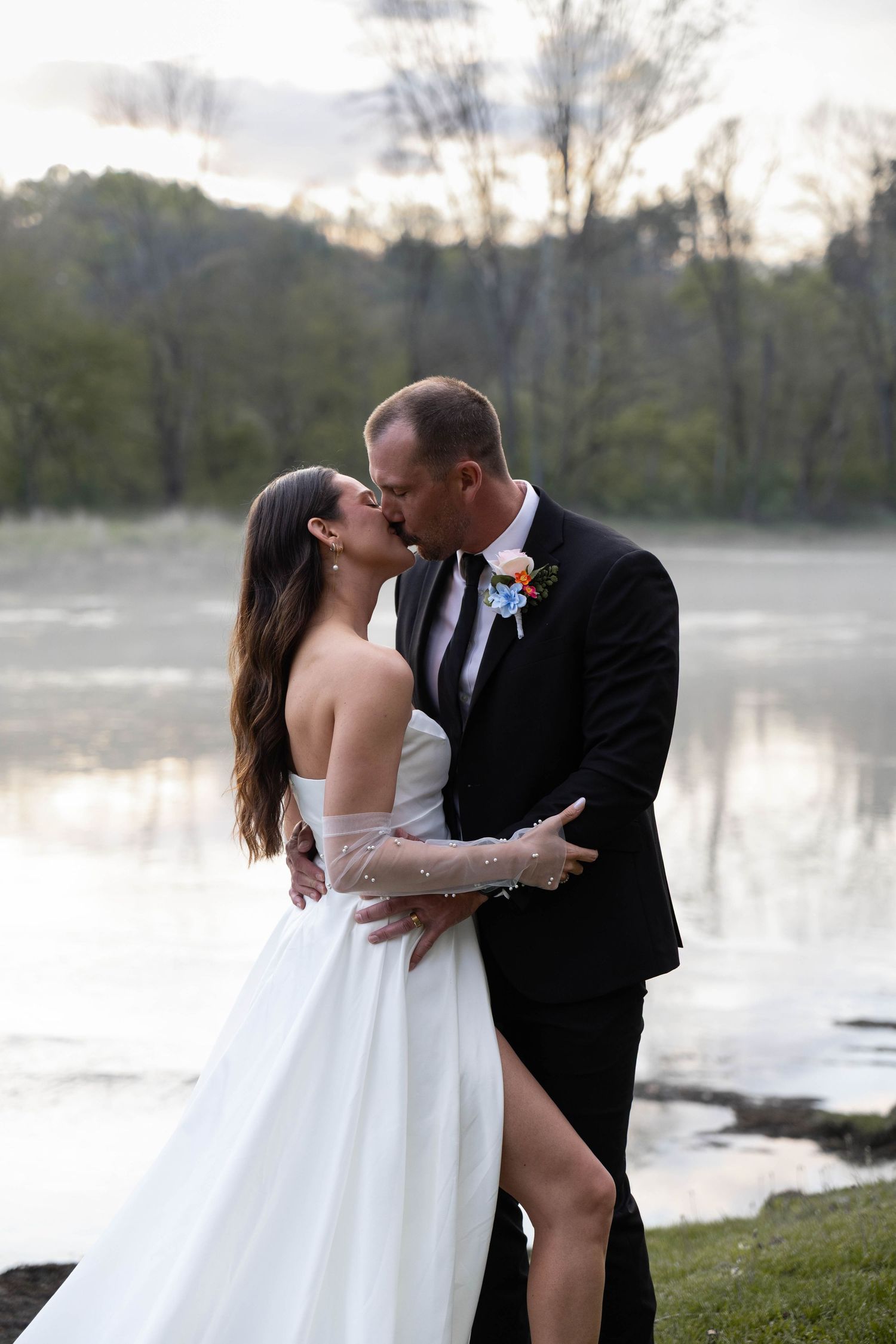 Bride and groom share a romantic kiss by a serene lake at sunset, she in a white gown, he in a black tuxedo.
