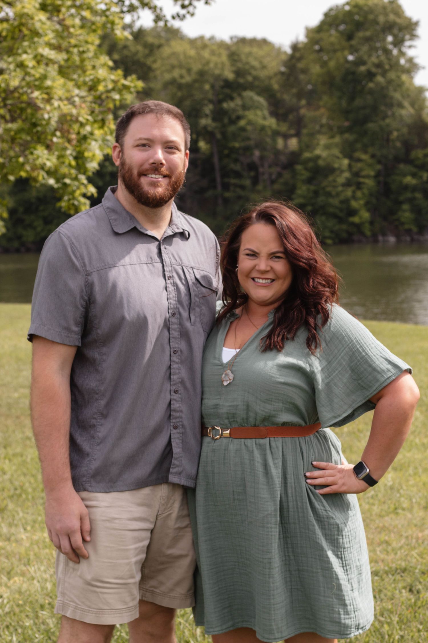 A couple stands together outdoors near a lake on a sunny day, wearing casual summer attire.