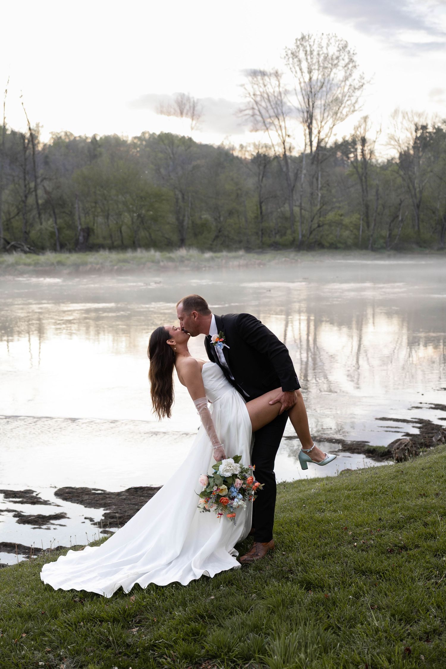 Groom dips bride for a romantic kiss by a misty river, bride holding colorful bouquet in white gown.