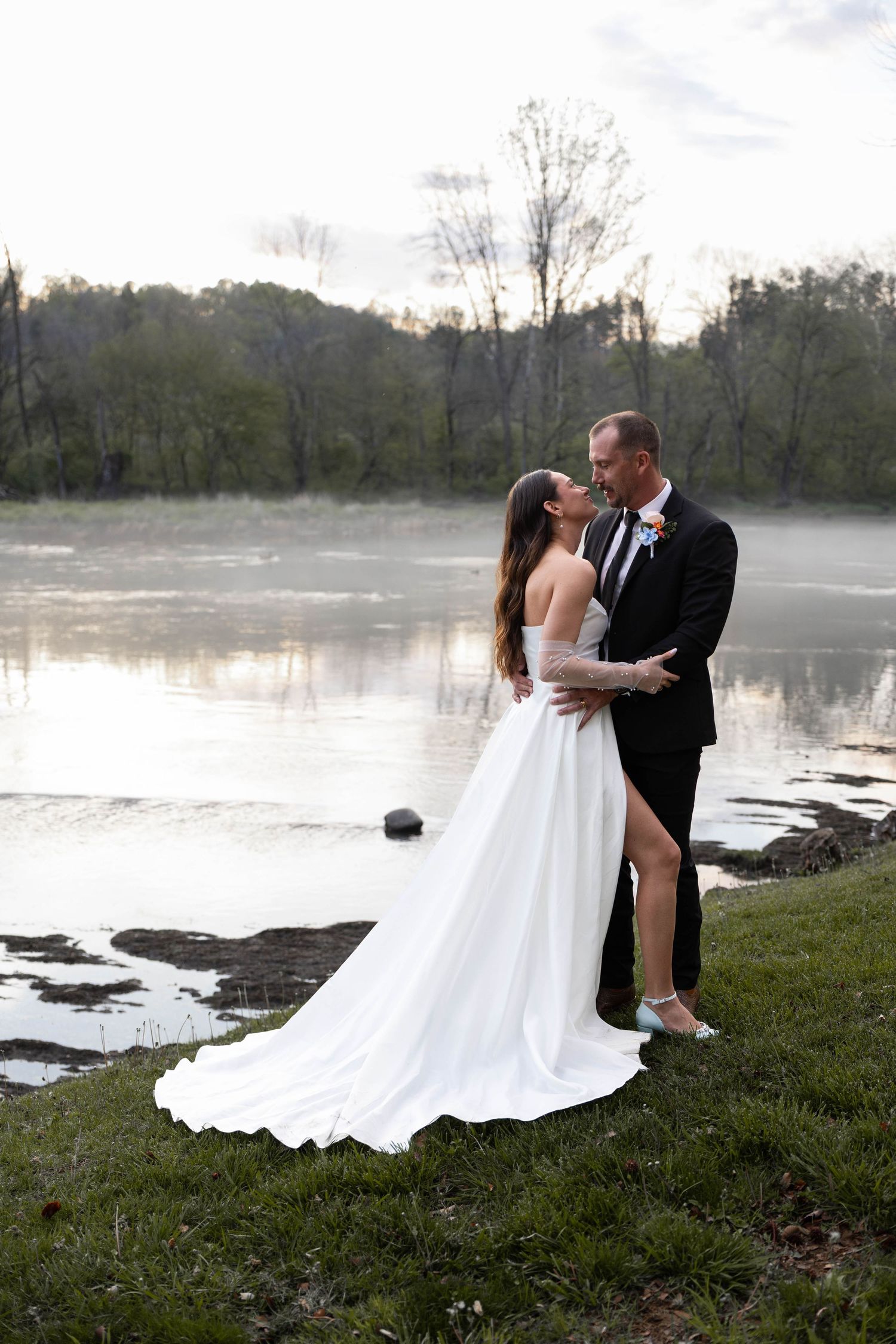 Bride and groom share a romantic moment by a misty lake, she in a flowing white gown with a long train.