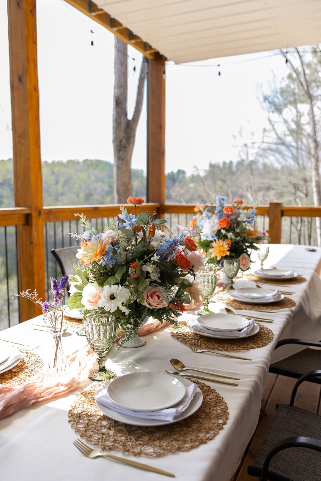 Colorful spring floral centerpiece on a rustic outdoor wedding reception table with gold cutlery and green goblets.