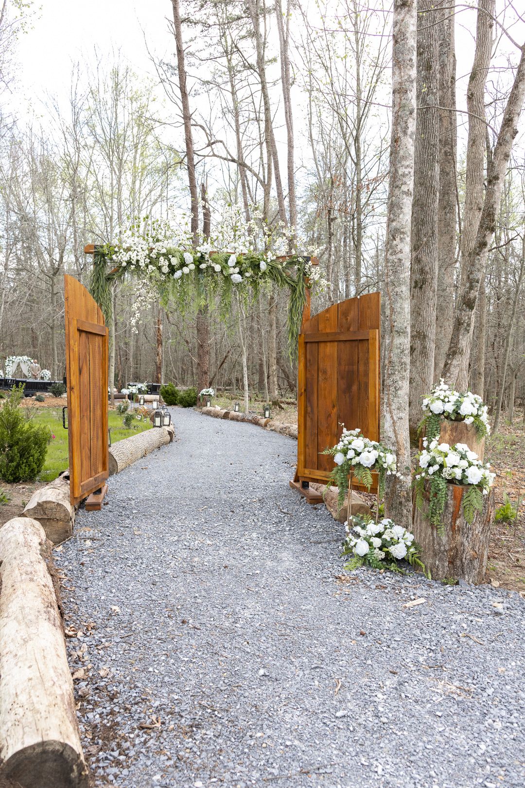 Rustic wooden wedding arch adorned with white flowers on a gravel path surrounded by trees.