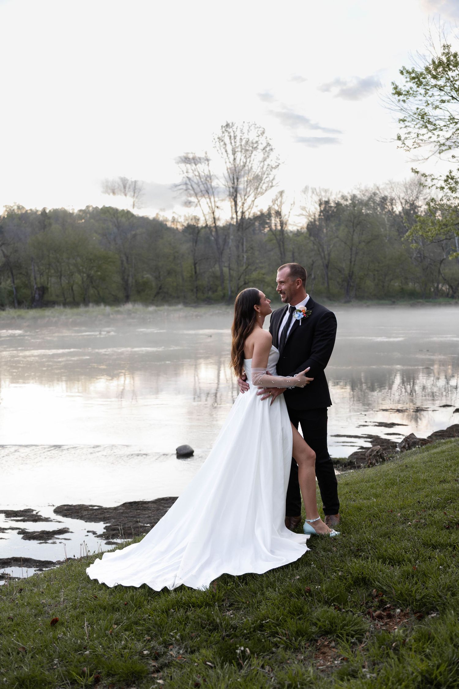 Wedding couple embraces near a foggy riverside at dusk, bride in white gown with slit, groom in black suit.