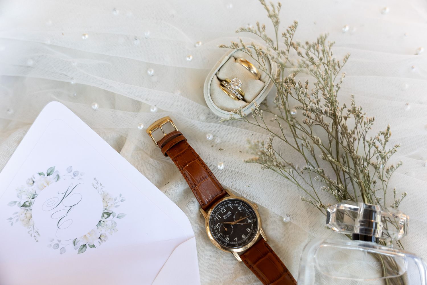 Elegant wedding flat lay with a brown leather watch, gold rings, floral invitation, pearl veil, and baby's breath bouquet.