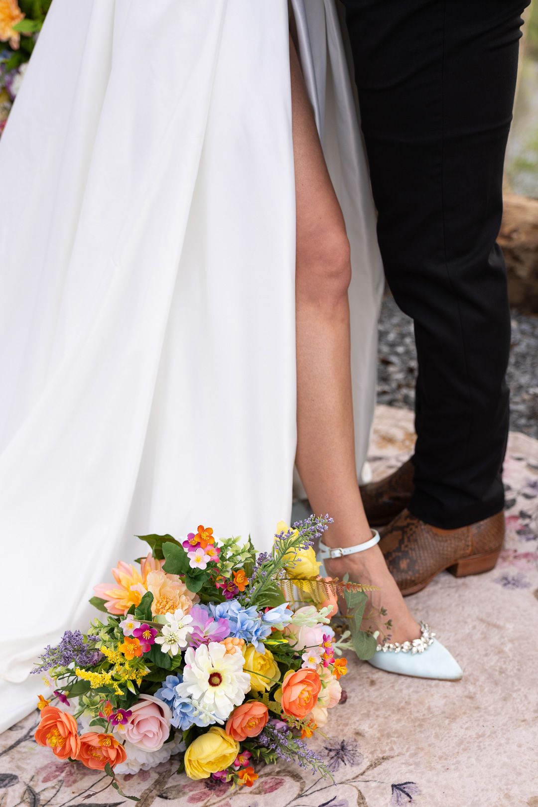 Colorful bridal bouquet with roses and wildflowers resting on ground beside bride in white gown and light blue pearl shoes.