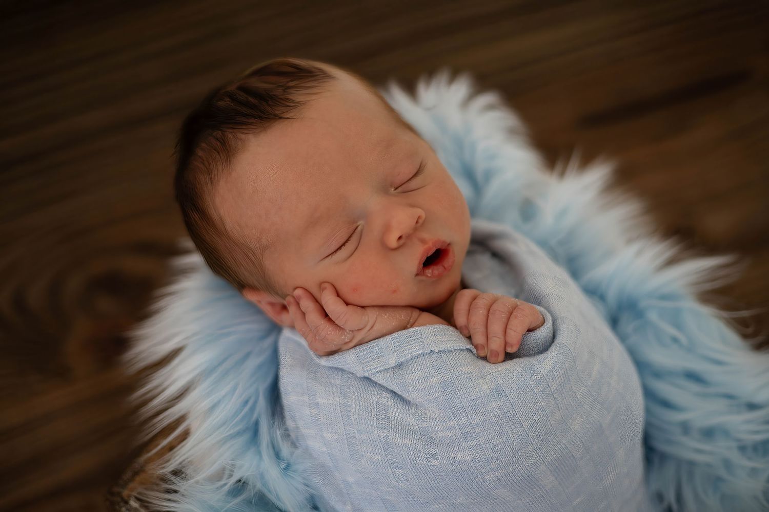 Sleeping newborn baby wrapped in gray swaddle on soft blue fur blanket during professional photography session.