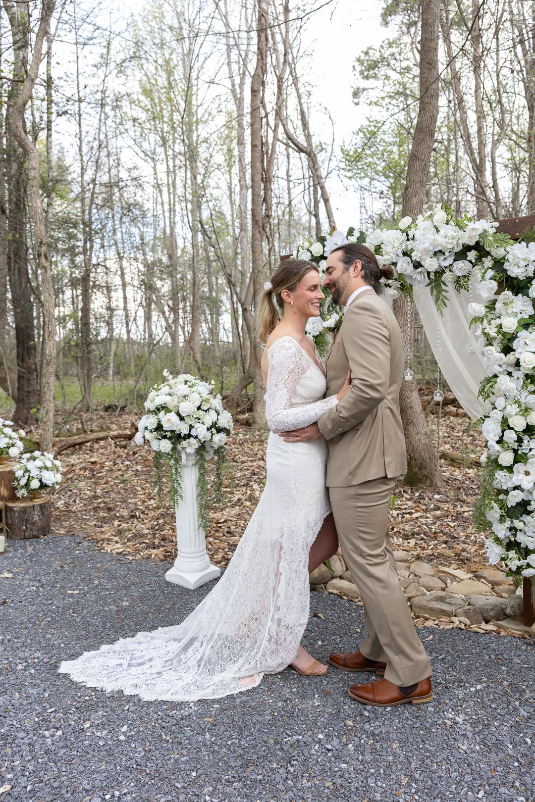 Bride and groom embrace outdoors at woodland wedding ceremony with white floral arch and flower arrangements.