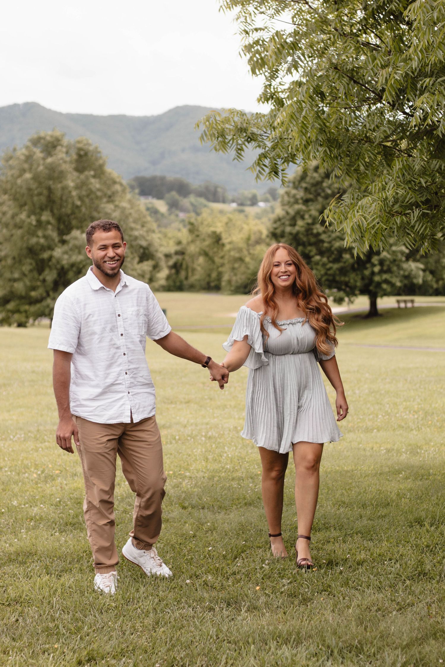 Couple walking hand in hand through a grassy field with mountains in background.