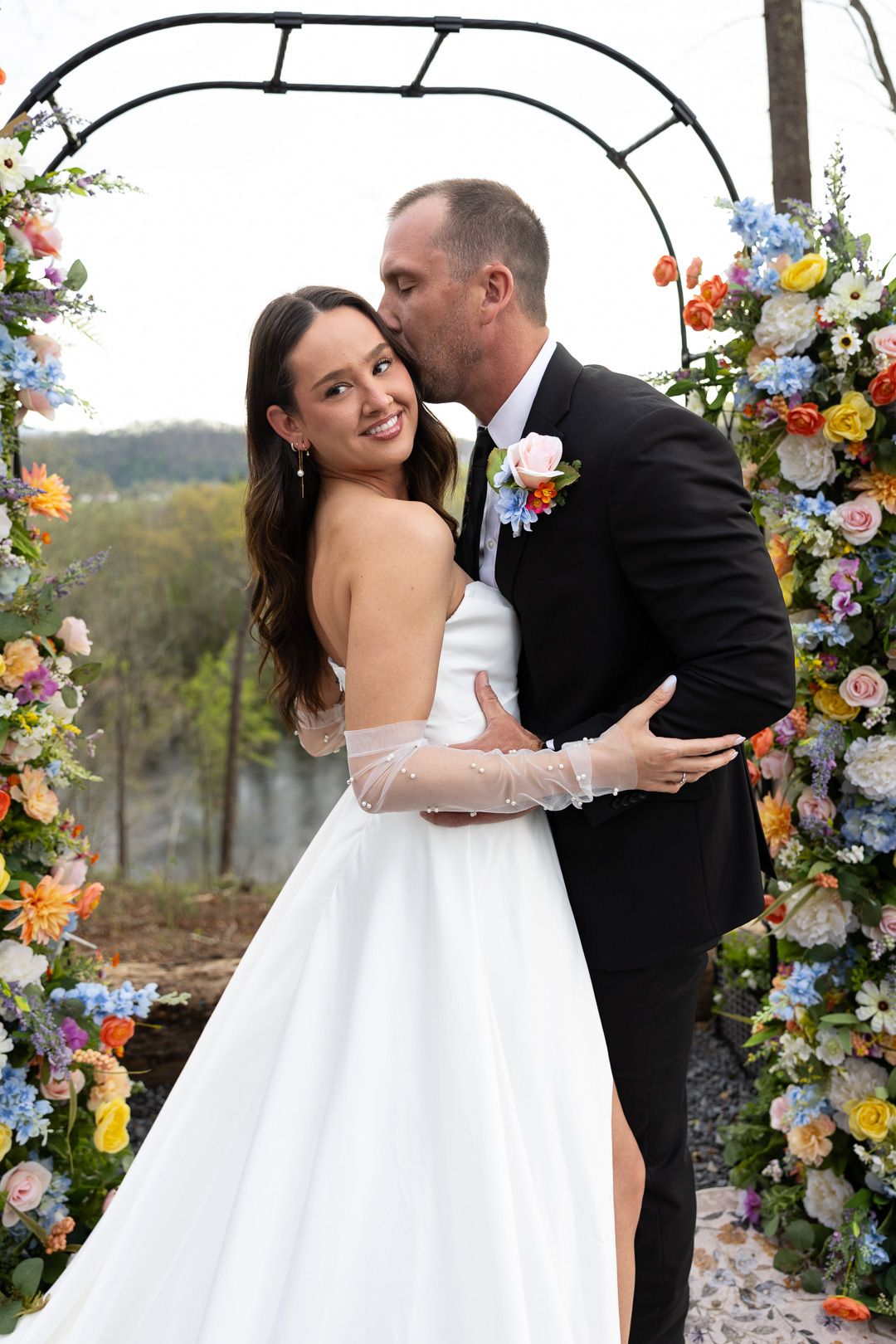 Bride and groom share a kiss under a colorful floral arch at an outdoor wedding ceremony.