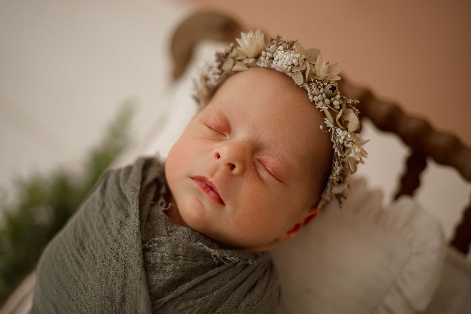Peaceful newborn in gray swaddle wearing dainty floral crown against neutral backdrop.