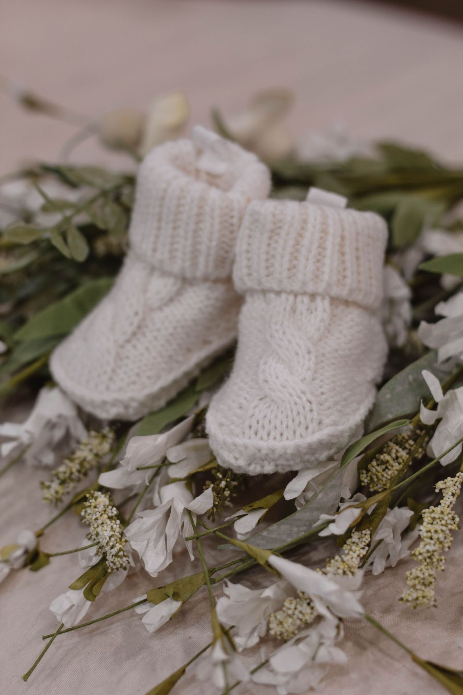Delicate white knitted baby booties arranged with white flowers and greenery.
