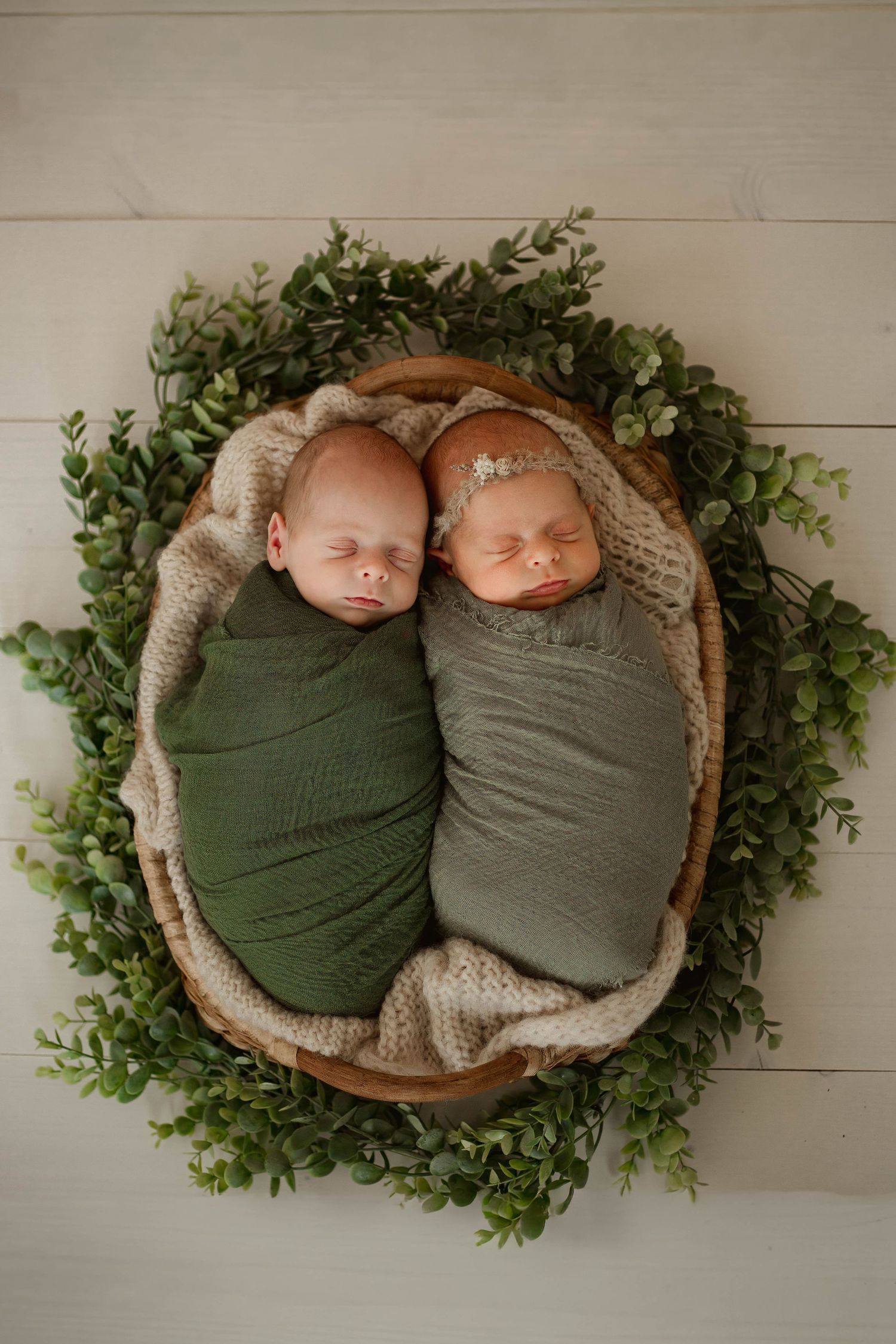 Two newborn babies swaddled in green and gray wraps lie side by side in wooden bowls surrounded by greenery.