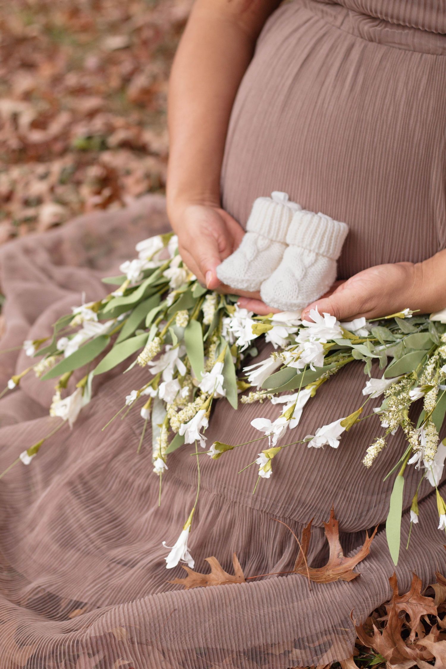 White baby booties and fresh flowers resting on a brown dress during maternity photoshoot.