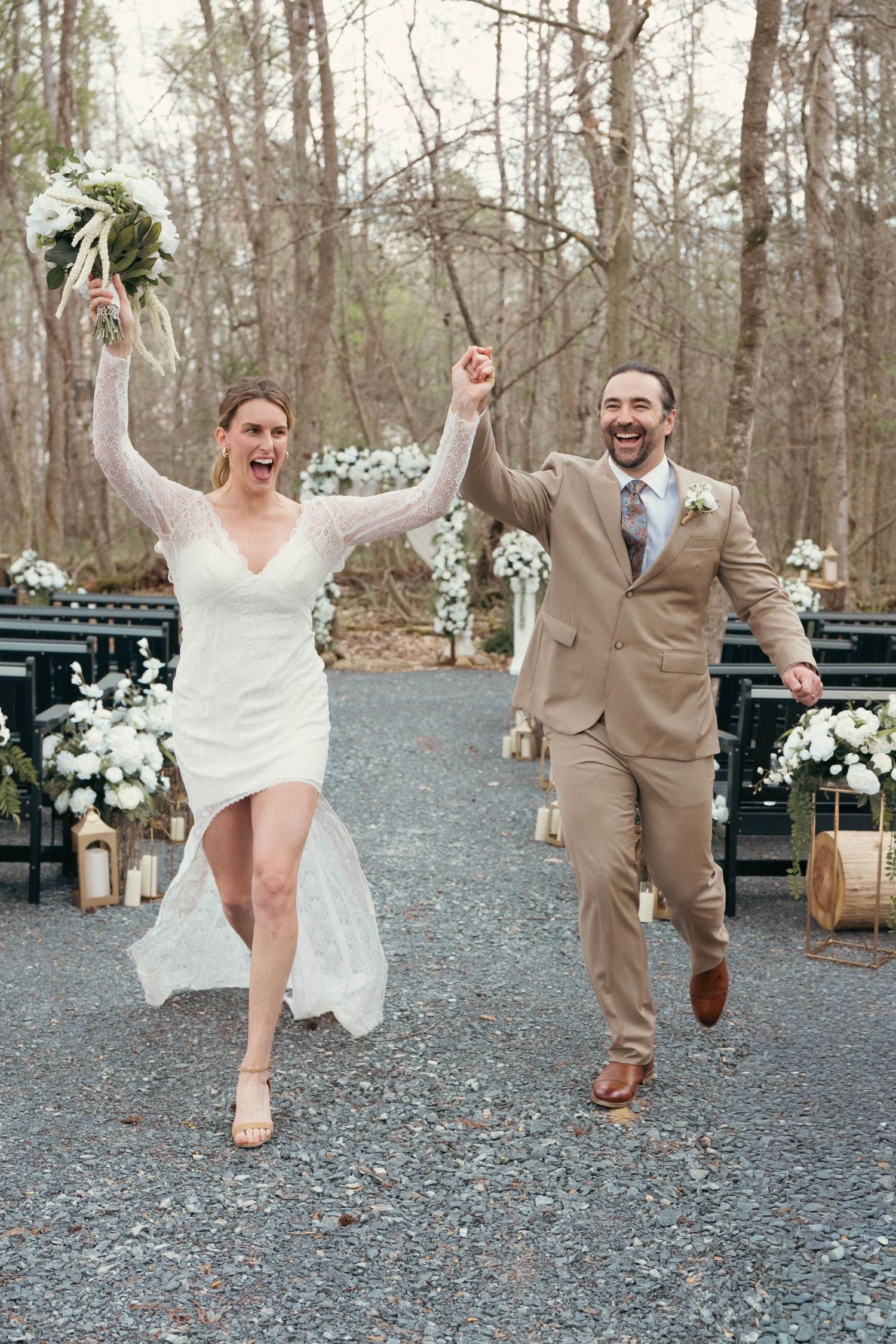 Joyful newlyweds celebrate after wedding ceremony, bride in white dress holding bouquet, groom in tan suit, outdoor woodland venue.